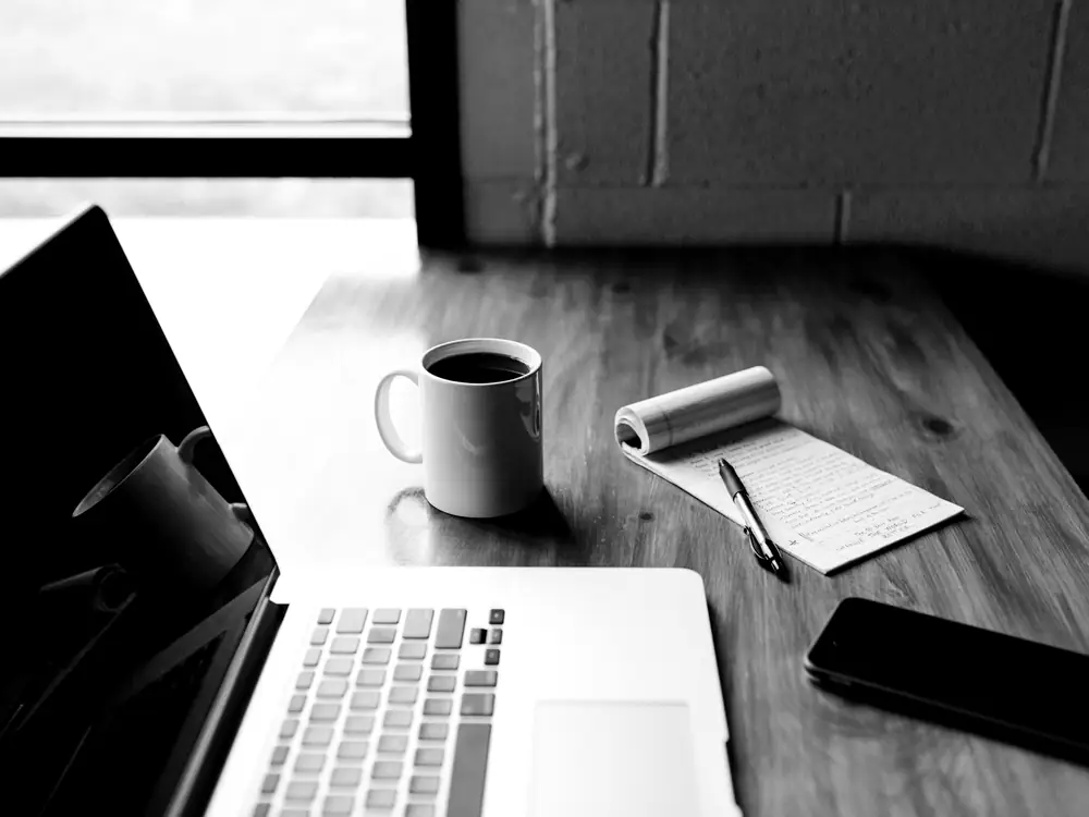 Portátil, cuaderno y taza de café sobre una mesa de madera junto a una ventana